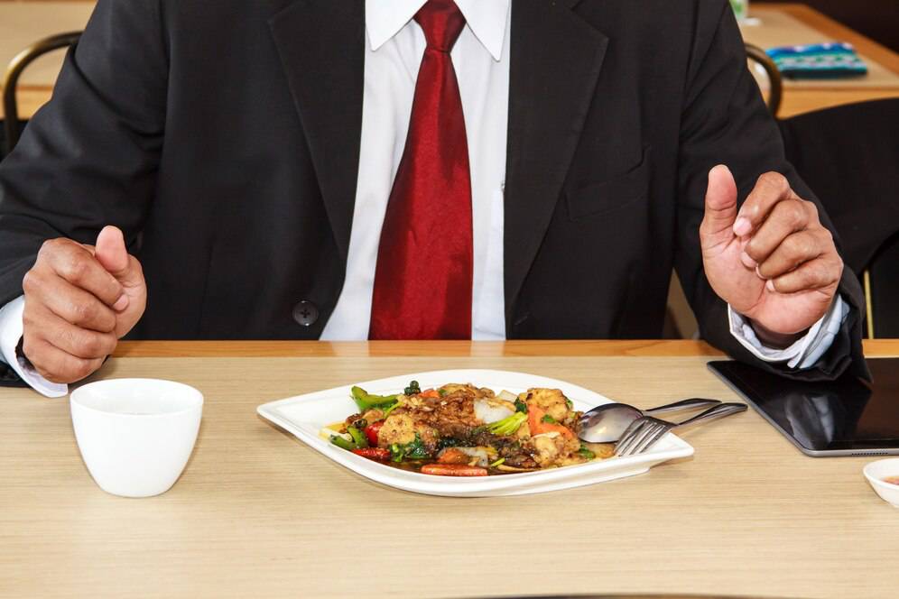 A businessperson at a table giving a thumbs-up gesture before a plate of food with a cup, fork, and smartphone nearby.