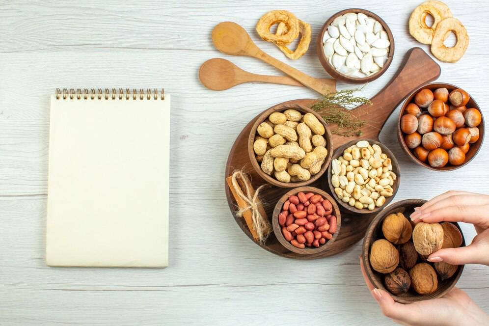 A variety of nuts in wooden bowls with a blank notebook and wooden spoons on a light wood surface.