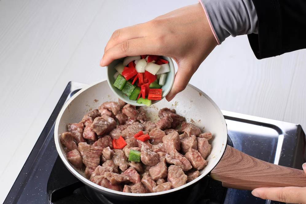 A person adding vegetables to sautéed meat in a frying pan on a stove.