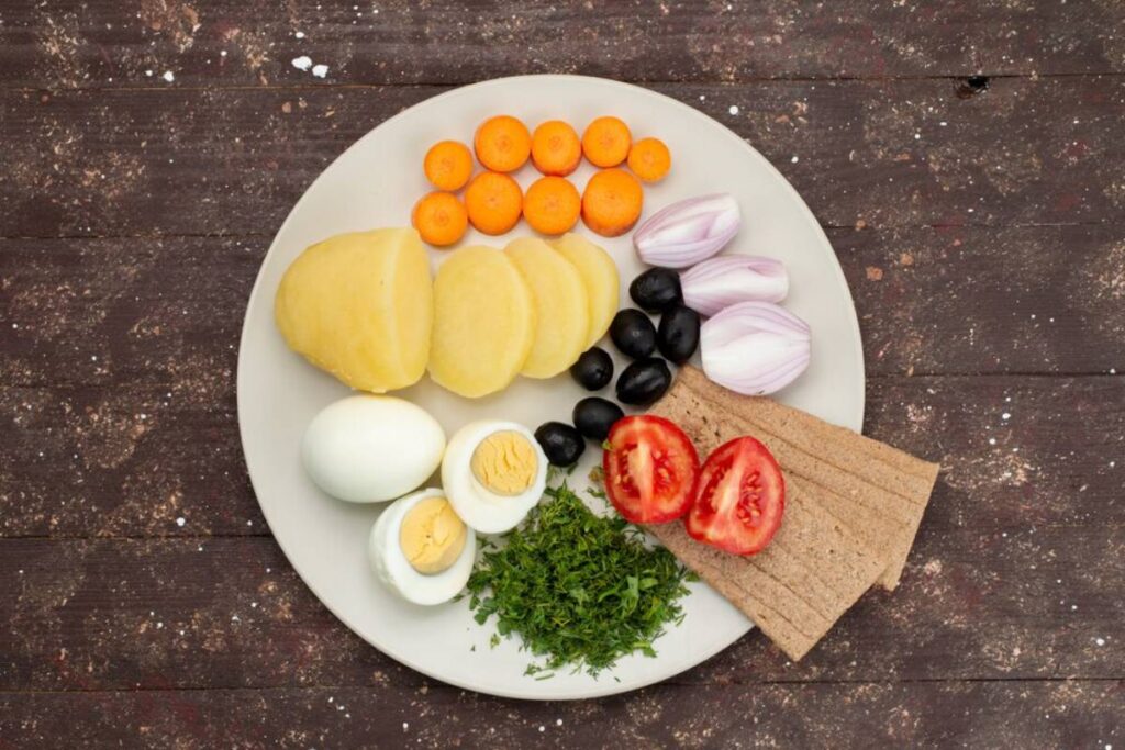 Plate of fresh ingredients including sliced potatoes, eggs, carrots, onions, black olives, tomatoes, herbs, and rye bread on a wooden background.