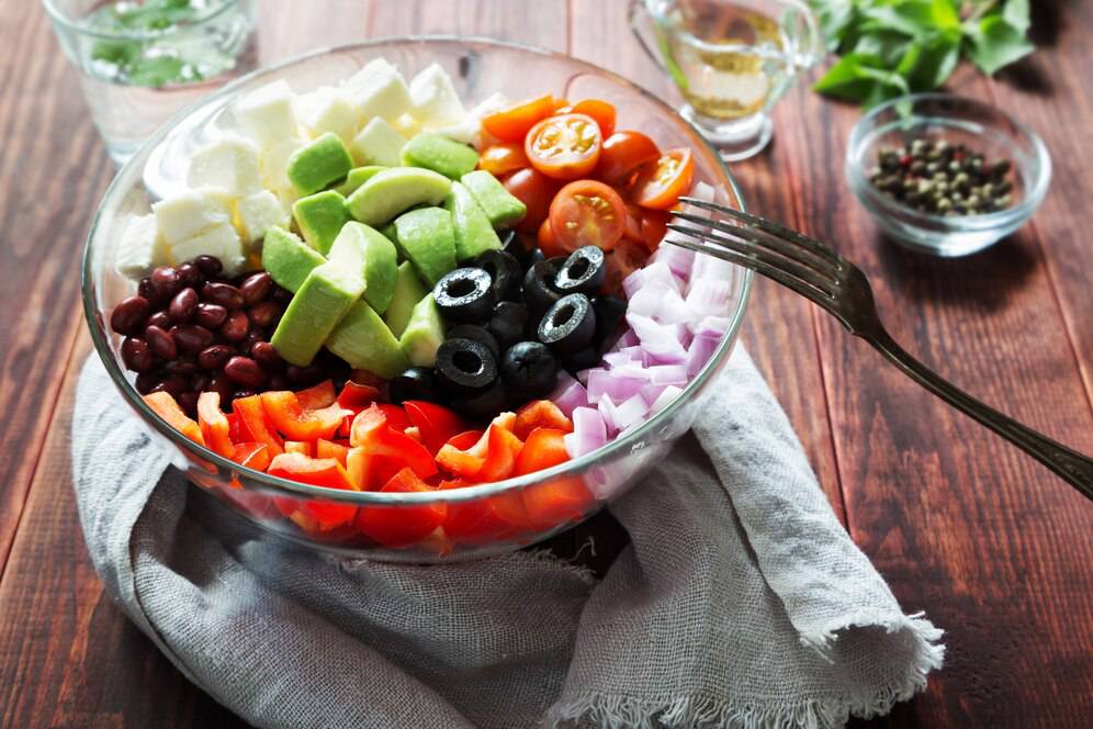 A colourful salad with beans, avocado, olives, cheese, onions, and tomatoes in a glass bowl on a wooden table.