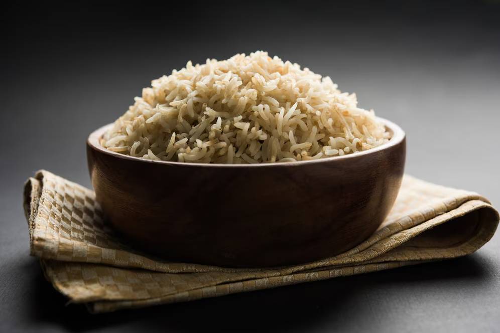 A bowl of cooked white rice on a woven mat, against a dark background.