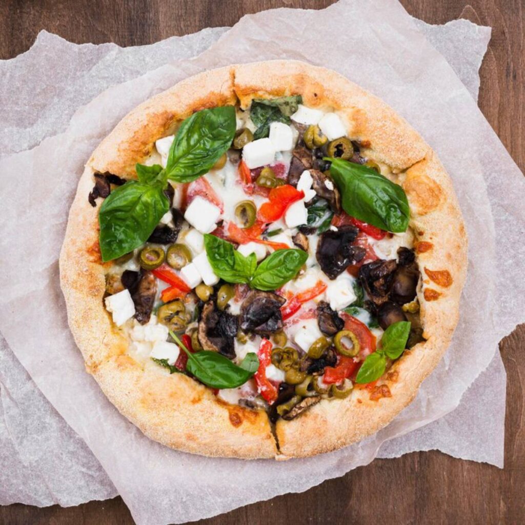 A freshly baked pizza topped with fresh basil, olives, red peppers, and feta cheese on a rustic wooden surface.