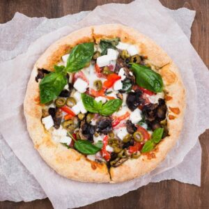 A freshly baked pizza topped with fresh basil, olives, red peppers, and feta cheese on a rustic wooden surface.