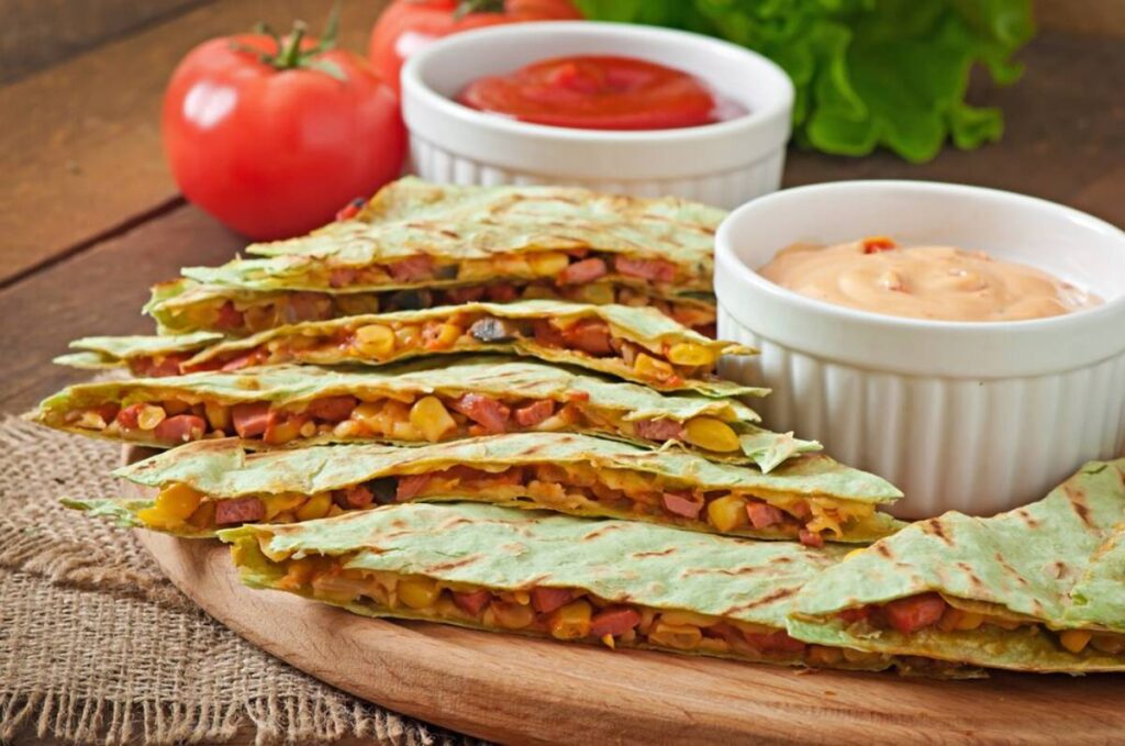Sliced vegetable quesadillas with dipping sauces in white bowls, garnished with fresh tomatoes and lettuce on a wooden table.