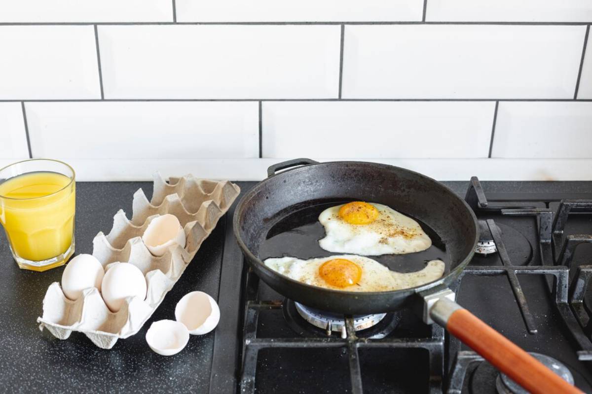 Fried eggs cooking in a skillet on a stove, with a glass of orange juice and egg carton nearby.