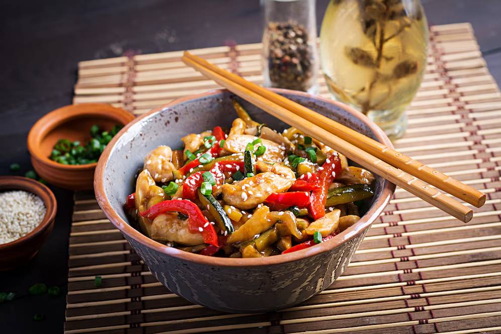 A bowl of stir-fried chicken with vegetables and sesame seeds, served with chopsticks on a bamboo mat.