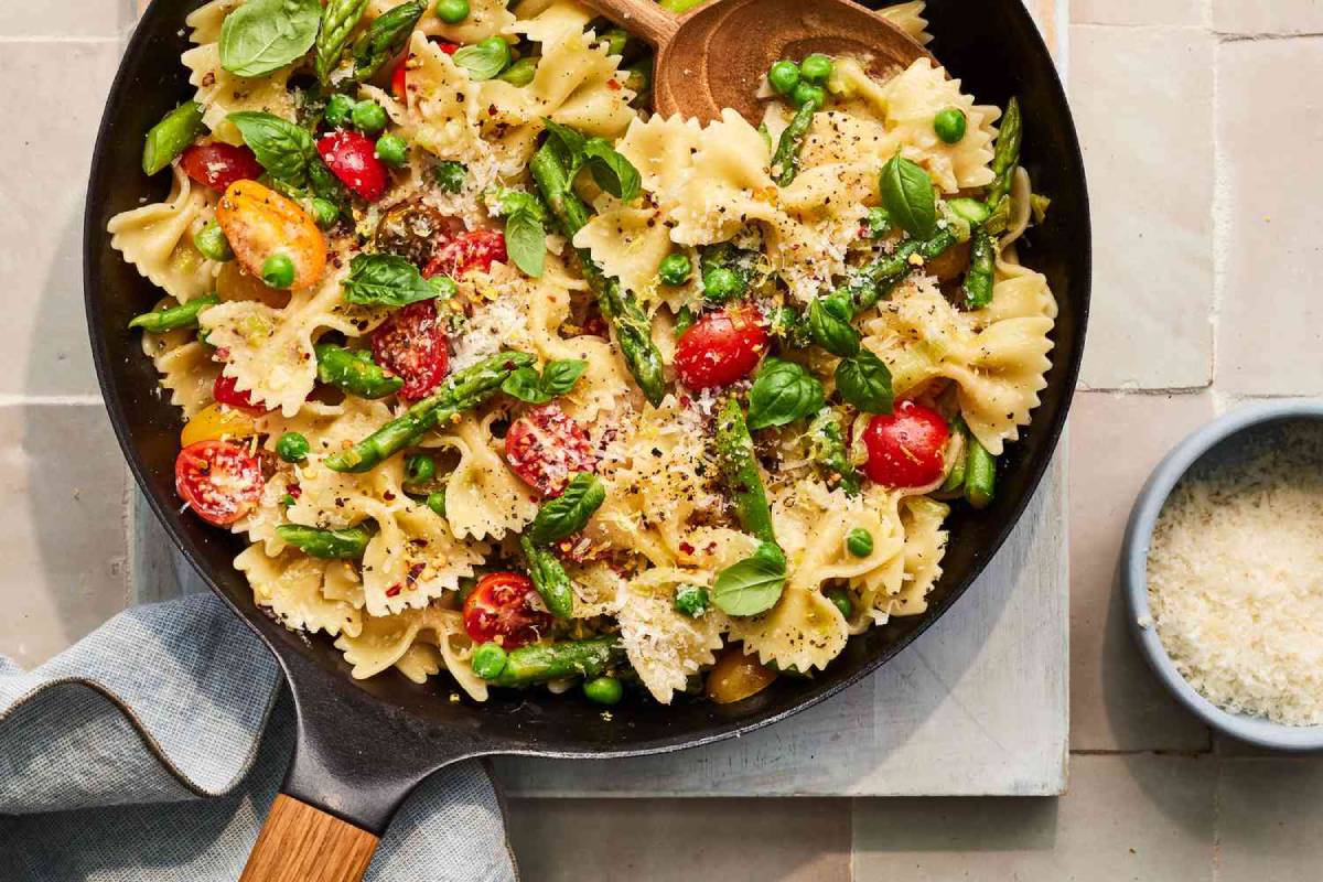 A vibrant farfalle pasta dish with asparagus, peas, cherry tomatoes, basil, and grated cheese, served in a skillet.