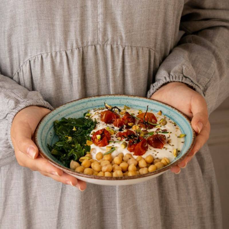 Person holding a bowl of chickpeas, roasted tomatoes, and greens, garnished with yogurt and seeds.