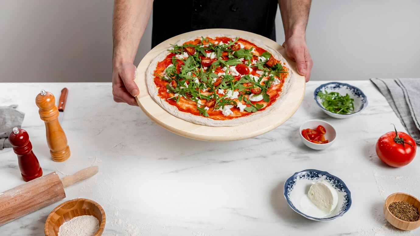 A chef holds a freshly topped pizza with tomatoes and arugula on a wooden pizza peel, surrounded by ingredients on a marble countertop.