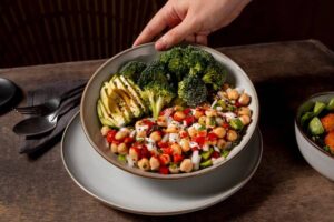 A hand serves a colorful bowl of salad with avocado, chickpeas, broccoli, and dressing on a dark wooden table.