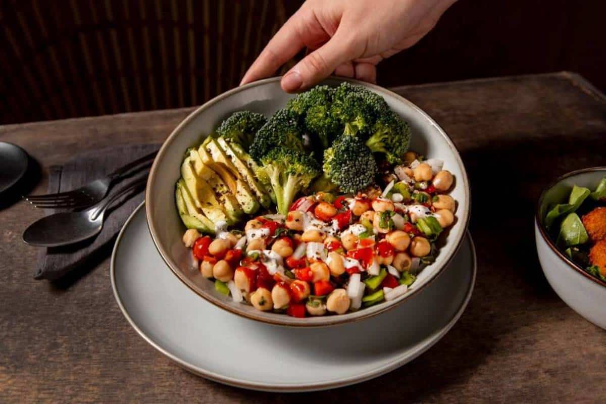 A hand serves a colorful bowl of salad with avocado, chickpeas, broccoli, and dressing on a dark wooden table.