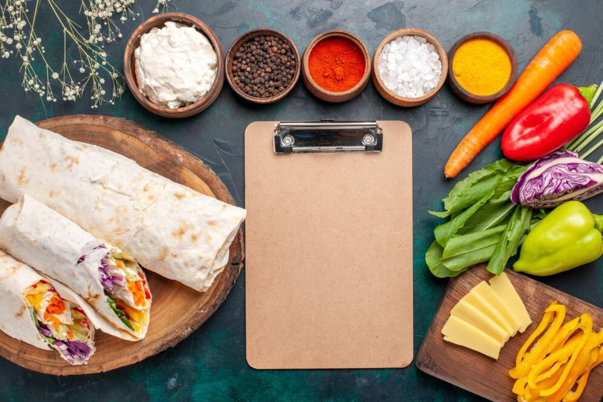 Top view of a meal preparation with fresh vegetables, spices, cheese, wraps, and a clipboard on a textured surface.