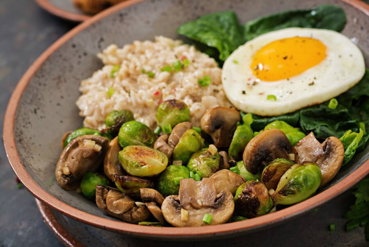 A healthy meal featuring brown rice, sautéed Brussels sprouts, mushrooms, spinach, and a fried egg in a bowl.