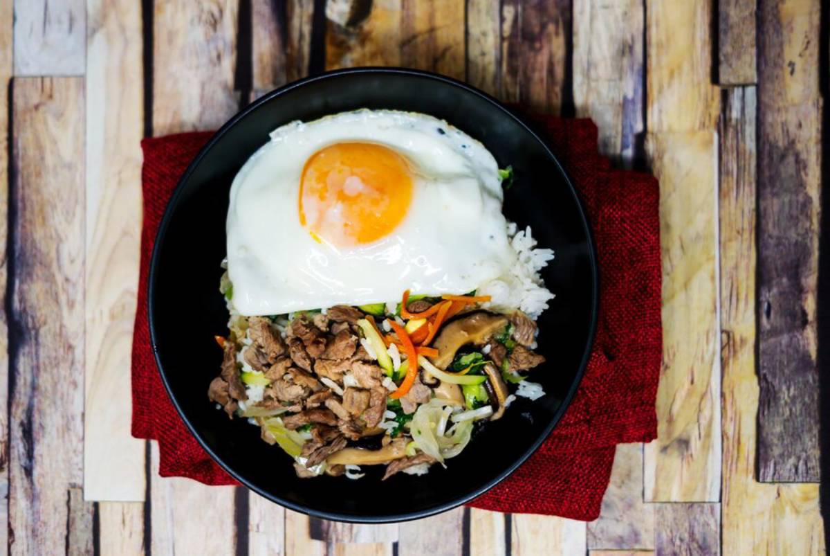 Rice topped with stir-fried beef, vegetables, and a sunny-side-up egg in a black bowl on a wooden table.