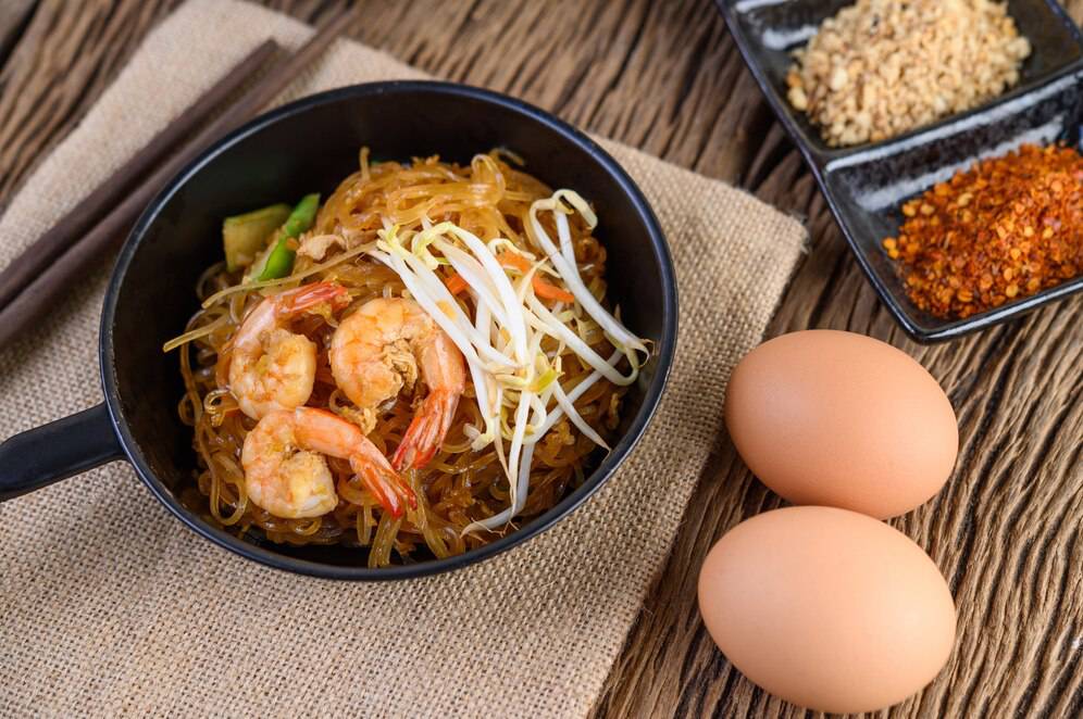 A black bowl of shrimp noodles topped with bean sprouts, accompanied by two eggs and seasoning on a wooden table.