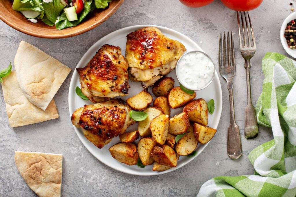 Grilled chicken thighs with roasted potatoes, fresh salad, pita bread, and a side of creamy dip on a gray background.