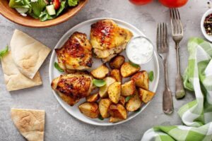Grilled chicken thighs with roasted potatoes, fresh salad, pita bread, and a side of creamy dip on a gray background.