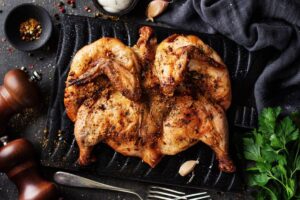 Roasted spatchcock chicken seasoned with herbs and spices on a dark grill pan, surrounded by garlic and cooking utensils.