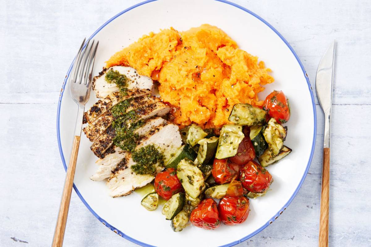 A plate with roasted chicken leg, creamy sweet potato mash, and sautéed cauliflower garnished with fresh herbs on a rustic wooden table.