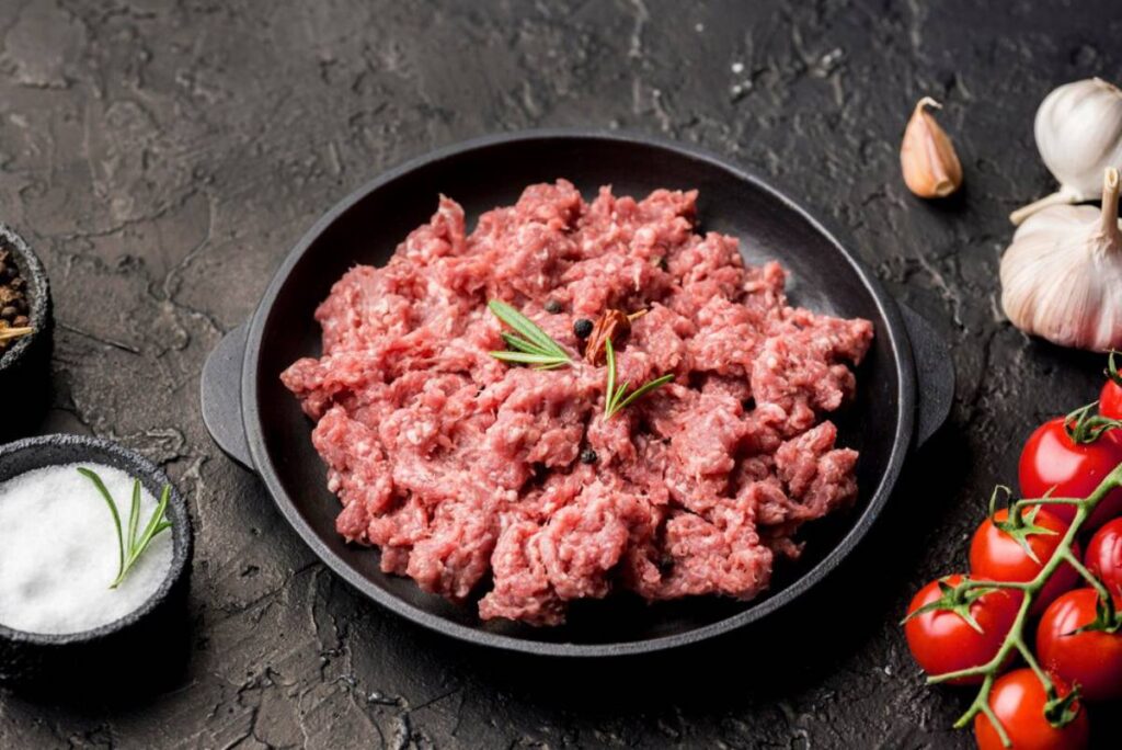 Raw minced meat in a cast iron skillet, seasoned with rosemary, alongside tomatoes and garlic on a dark surface.