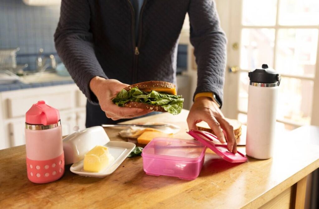 Person preparing a sandwich with fresh lettuce and cheese in a bright kitchen, placing it in a pink lunchbox.
