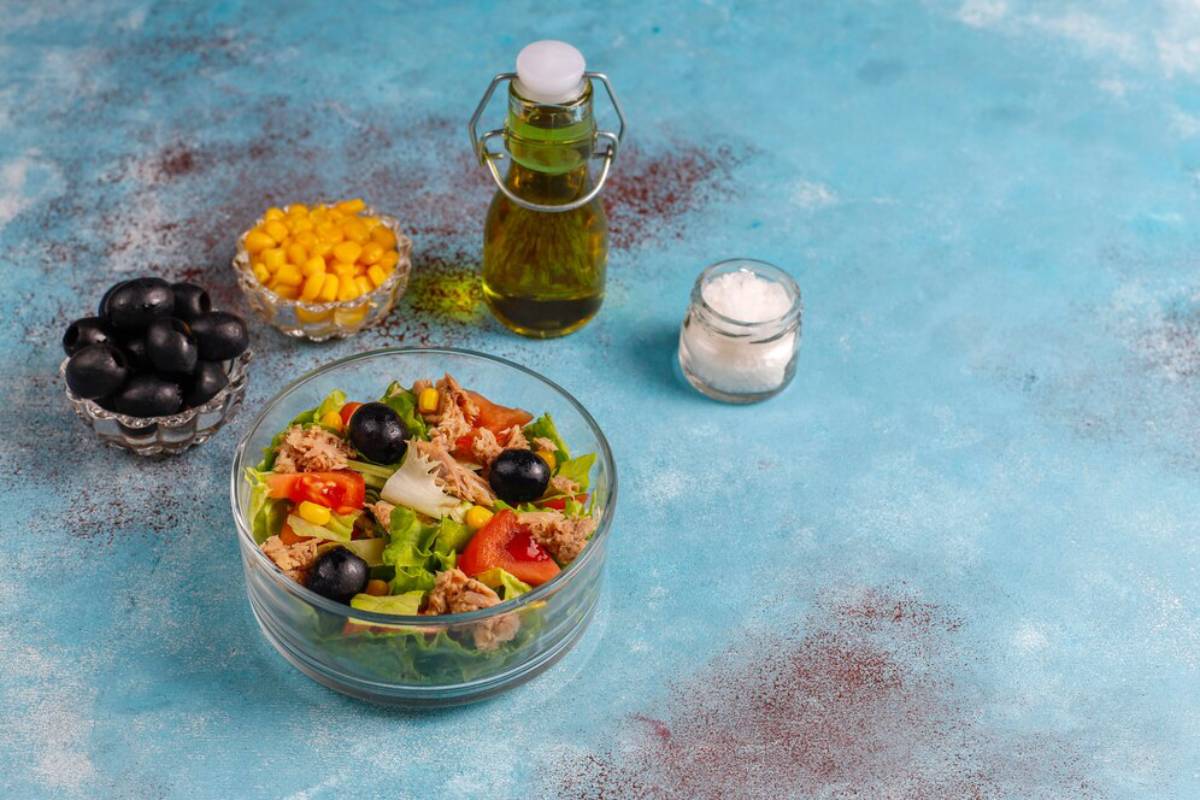 A glass bowl of salad with tuna, olives, and vegetables next to condiments on a blue background.