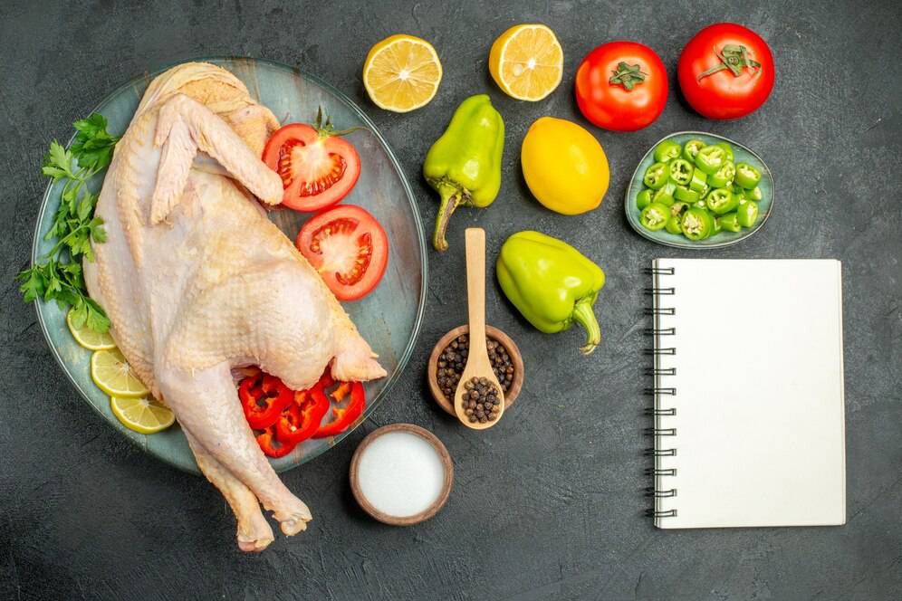 Raw chicken with tomatoes, lemons, herbs, peppers, salt, and a blank notebook on a dark surface.