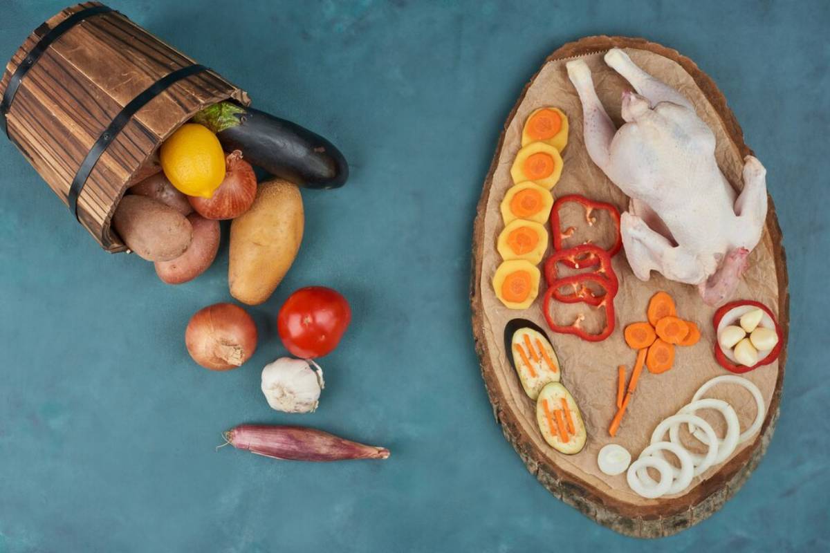 Various fresh vegetables spilled from a basket and a raw chicken with sliced vegetables on a wooden board.