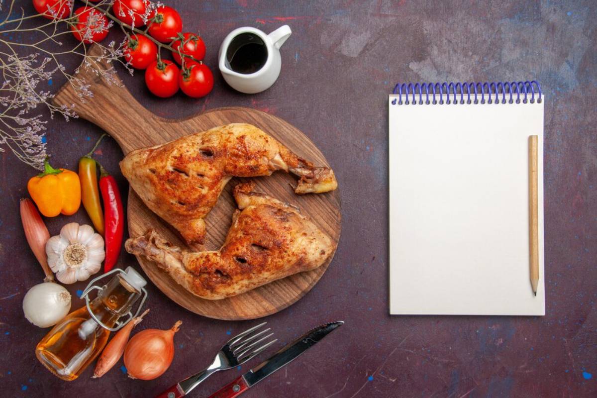 Grilled chicken legs on a wooden board with vegetables, sauce, and a blank notepad on a textured surface.
