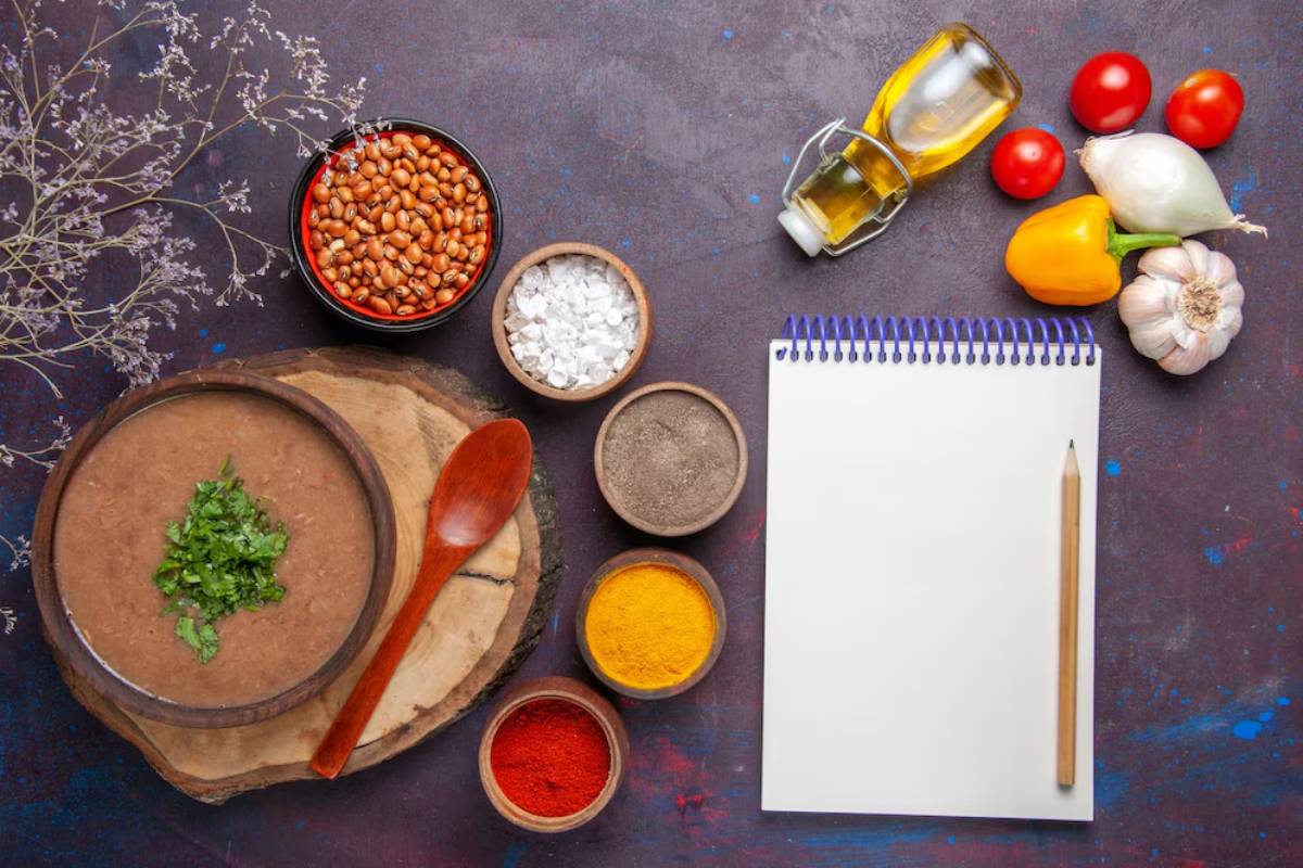 Ingredients for a recipe displayed including beans, olive oil, tomatoes, garlic, spices, a bowl of soup garnished with herbs, and a blank notebook.