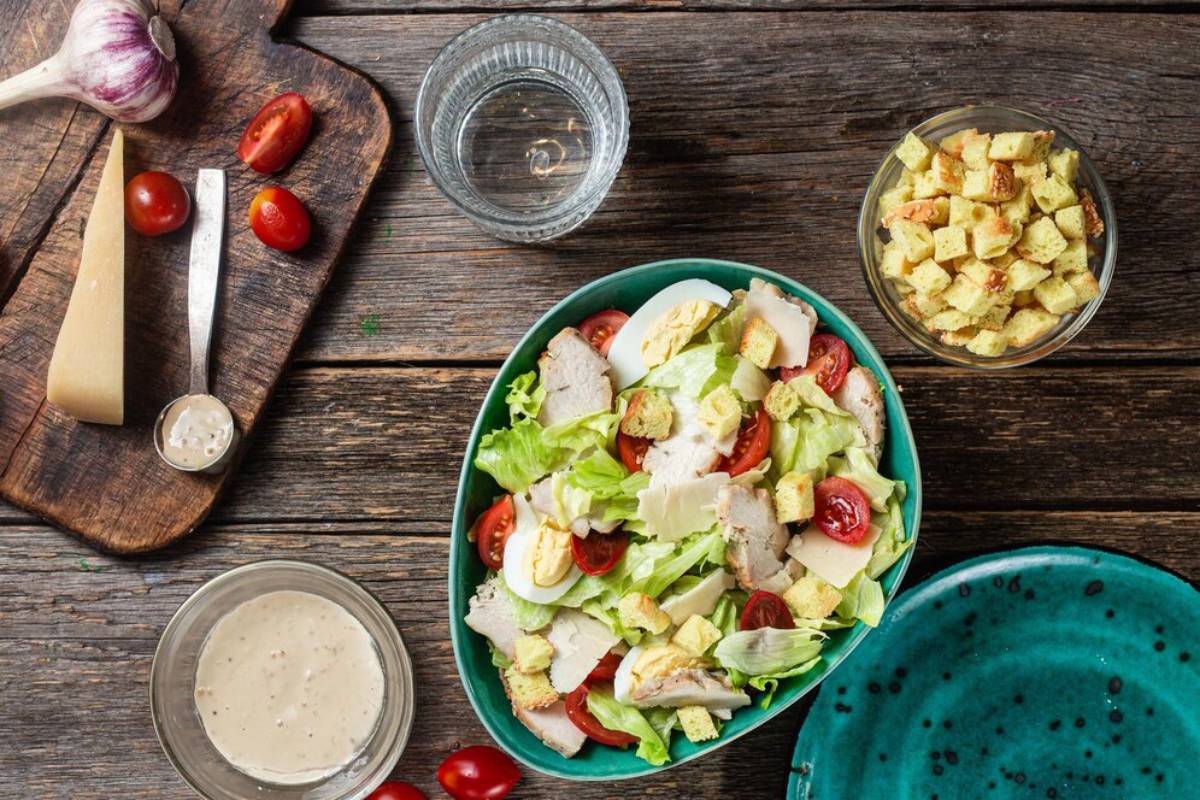 A fresh Caesar salad with chicken, eggs, tomatoes, and croutons, served on a wooden table with dressing and ingredients nearby.