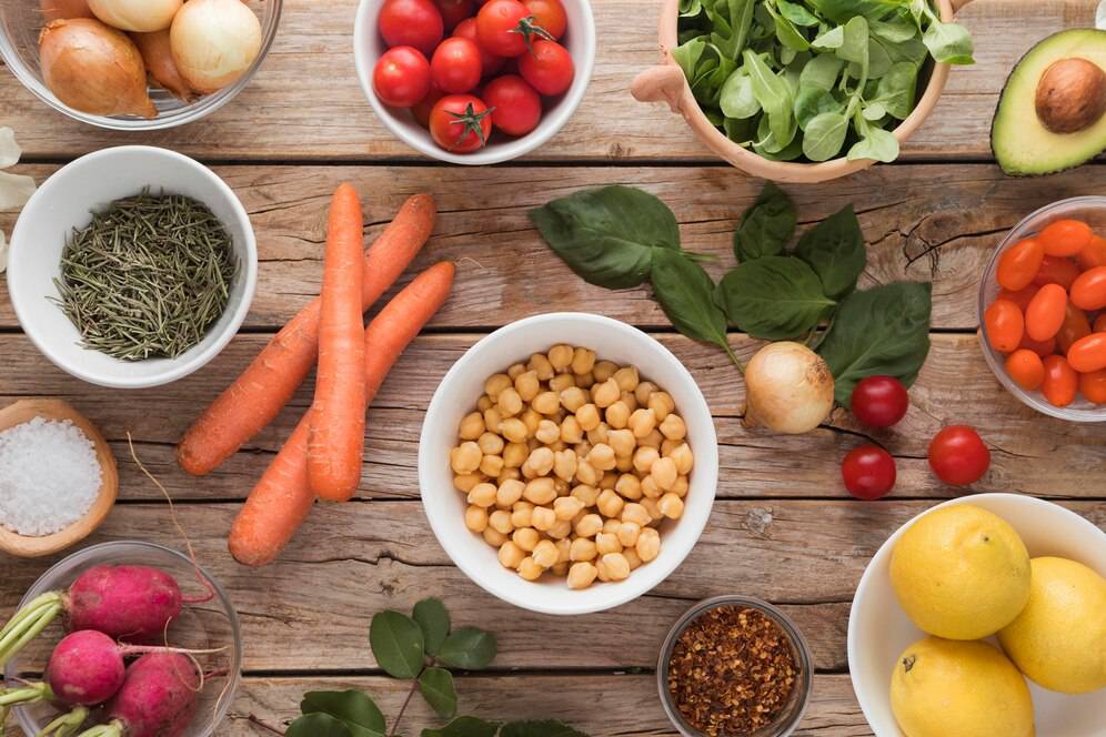 Assorted fresh vegetables and legumes on a wooden table, including carrots, tomatoes, chickpeas, and avocados.