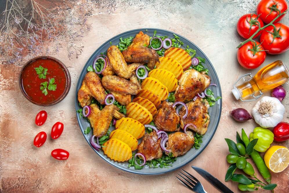 Plate of roasted chicken wings, sliced potatoes, and fresh vegetables, with sauce and a mug of beer on a textured table.