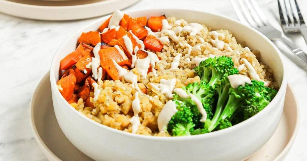 A bowl of quinoa with roasted carrots, broccoli, and a drizzle of white sauce.