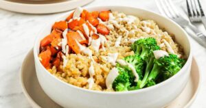 A bowl of quinoa with roasted carrots, broccoli, and a drizzle of white sauce.