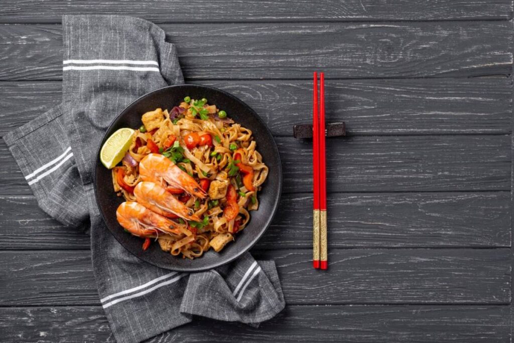A plate of stir-fried noodles with shrimp, vegetables, and a lime wedge, served with red chopsticks on a dark wooden table.