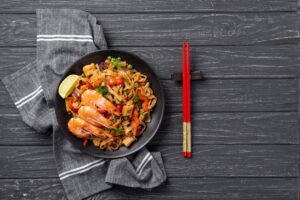 A plate of stir-fried noodles with shrimp, vegetables, and a lime wedge, served with red chopsticks on a dark wooden table.