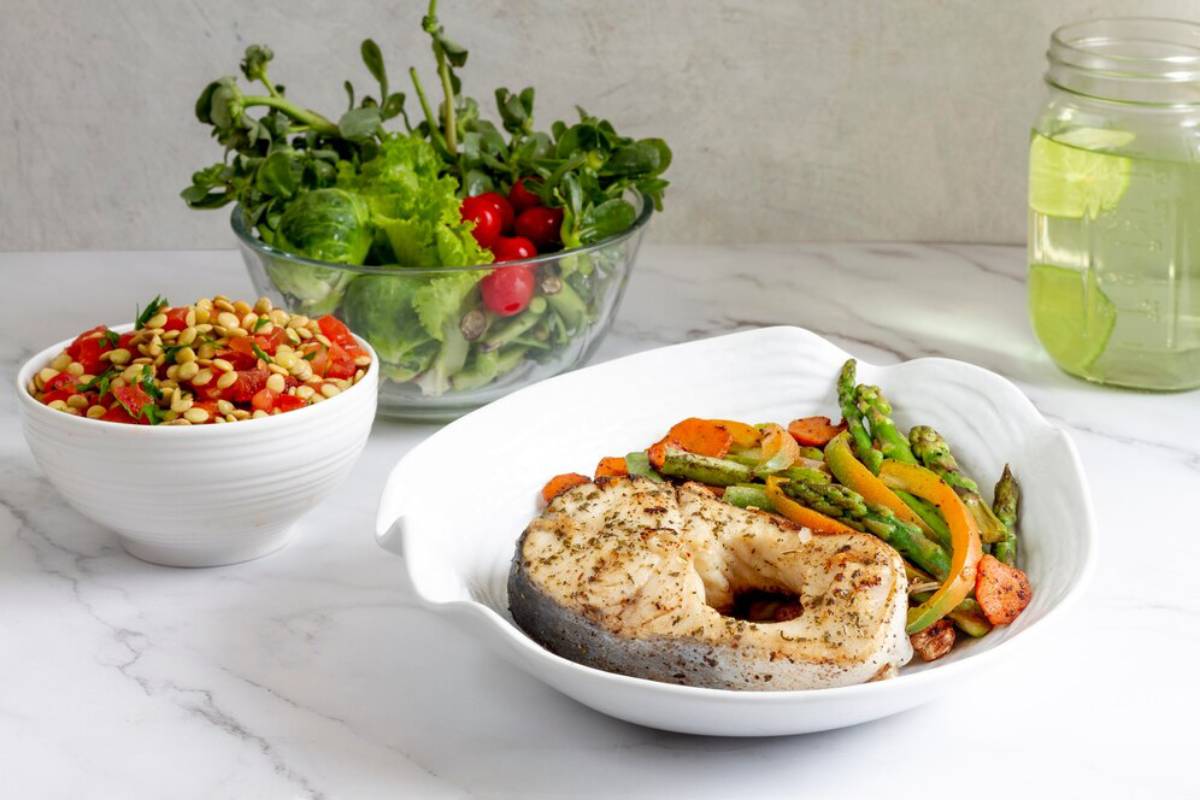Grilled fish with mixed vegetables, a side of green salad, tomato pine nut salad, and a jar of lime water on a marble table.