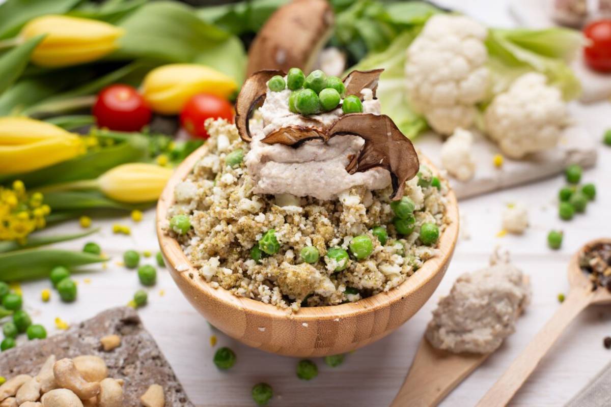 Bowl of couscous with peas, roasted mushrooms, and a creamy sauce, surrounded by fresh vegetables.