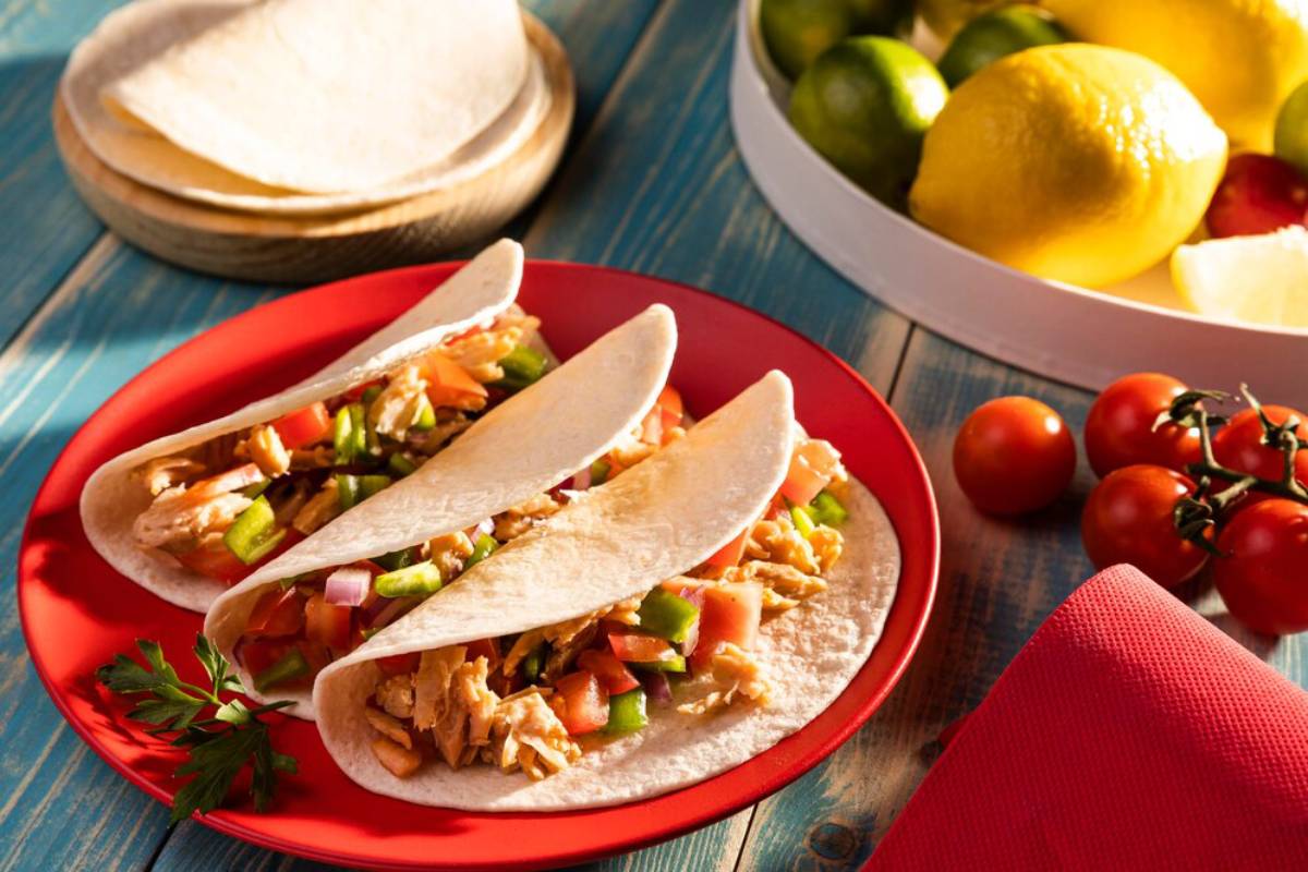 Two chicken tacos on a red plate with vegetables and tortillas in the background.