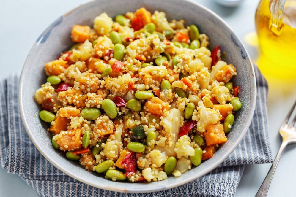 Bowl of quinoa salad with mixed vegetables, including carrots, edamame, and red peppers, served on a blue-striped napkin.