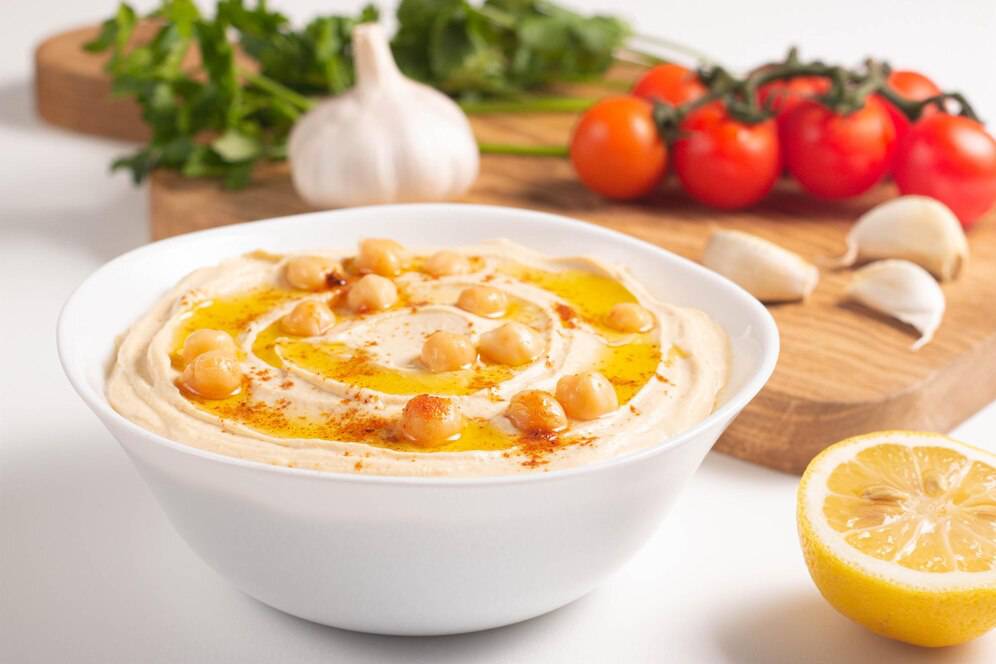 Bowl of hummus topped with chickpeas, olive oil, and paprika, surrounded by fresh ingredients like tomatoes, garlic, and lemon.