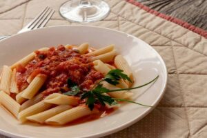 A bowl of penne pasta topped with tomato sauce and a sprig of parsley, served on a quilted mat.