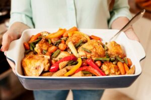 Person holding a baking dish with roasted chicken and colorful mixed vegetables.