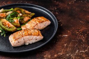 Two grilled salmon fillets with sautéed vegetables on a black plate, rustic dark background.