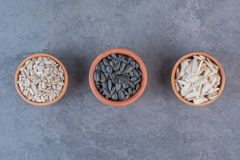 Three terracotta bowls containing sunflower seeds, pumpkin seeds, and squash seeds on a grey surface.