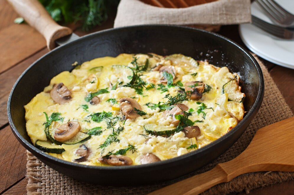A golden frittata with mushrooms, zucchini, and fresh herbs in a skillet, set on a rustic wooden table.
