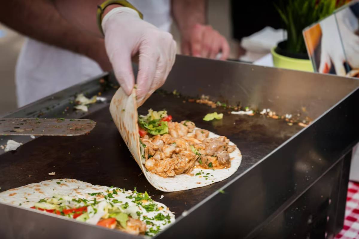 Person wearing gloves prepares chicken wraps on a hot plate at a food stall.