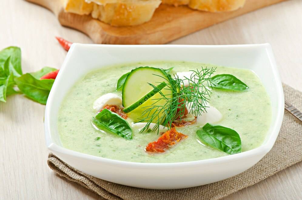 A bowl of creamy green soup garnished with cucumber, dill, sun-dried tomatoes, and basil leaves, served on a wooden table.
